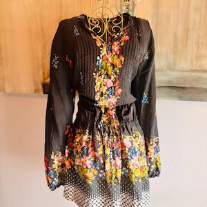 Puede incluir: Vestido negro con estampado floral y mangas largas. El vestido presenta un cuello en V con pliegues y una cintura con cord&oacute;n. La falda tiene un estampado floral con un dobladillo de lunares blancos. El vestido se exhibe en un maniqu&iacute; dorado.