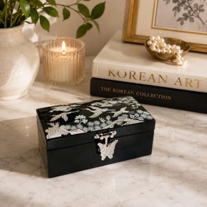 May include: A rectangular black lacquer jewelry box featuring an inlaid mother-of-pearl design of cranes and pine trees on the lid. The box is secured with a silver-toned metal butterfly-shaped latch on the front center of the lid.
