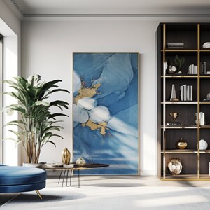May include: A blue and gold abstract painting hangs on a white wall in a modern living room. A blue chaise lounge sits in front of a round coffee table with gold accents. A tall bookshelf with gold accents and shelves filled with books and decorative objects is on the right side of the image.