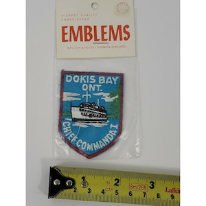 Op de afbeelding: Geborduurde patch met de tekst "DOKIS BAY ONT." en een bootillustratie. De patch is blauw met rode en witte accenten. De verpakking leest "EMBLEMS" en "HIGHEST QUALITY EMBROIDERED".