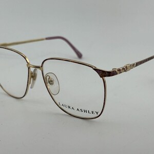 Könnte beinhalten: Goldfarbene Laura Ashley Brille mit rechteckigem Rahmen. Die Bügel und die oberen Rahmenecken haben ein dekoratives Muster. Die Bügel und die Rahmenarme sind dunkelburgunderfarben. Die Gläser sind klar.