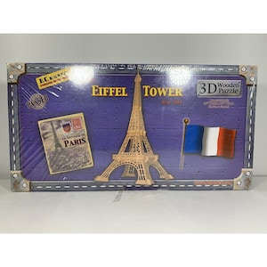May include: A boxed 3D wooden puzzle of the Eiffel Tower. The box features the Eiffel Tower, a French flag, and a vintage postcard. The text on the box reads "Eiffel Tower" and "Le Souvenir de Paris".