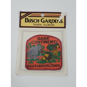 May include: A vintage Busch Gardens Tampa patch. The patch has an orange background with the words "DARK CONTINENT" and illustrations of an elephant, lion, and giraffe. It is in a clear plastic package.
