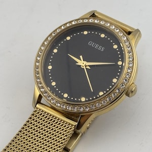 May include: A gold-coloured Guess watch with a black face and a mesh strap. The watch face is encircled with a row of small, clear crystals. The watch hands and the word "GUESS" are in gold.