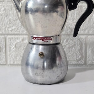 以下が含まれることがあります： 黒いプラスチック製のハンドルと蓋に黒いノブが付いた、銀色のアルミニウム製直火式エスプレッソメーカー。中央の帯にある赤いラベルには「Signora caffettiera」という文字が記されています。ベース部分には小さな真鍮製の圧力バルブが付いています。