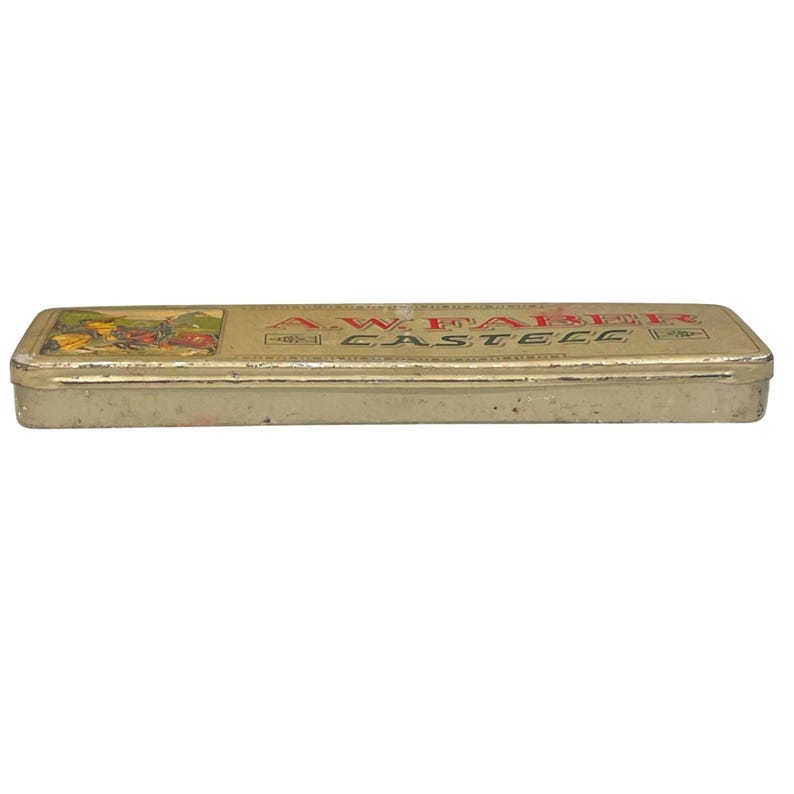 May include: A vintage rectangular pencil case with a gold finish. The case features the text "A.W. FABER CASTELL" in red lettering. A small illustration is on the left side of the case. The case is likely made of metal.