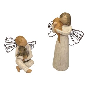 May include: Two angel figurines, one seated and one standing, each holding a small dog. The angels are dressed in white robes and have wire wings. The dogs are brown and gray. The figurines are made of a carved, textured material.