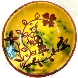 May include: A small, round ceramic bowl with a yellow interior and a brown rim. The bowl features a hand-painted design of a bird, flowers, and leaves in brown against a green and yellow background. The bowl is likely handmade.