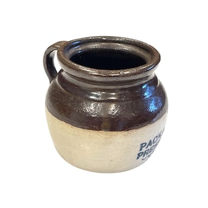 May include: A small, antique stoneware jar with a dark brown top and handle, and a cream-colored base. The jar has a cylindrical shape with a wide opening and a printed label in blue. The jar is likely used for preserving food.