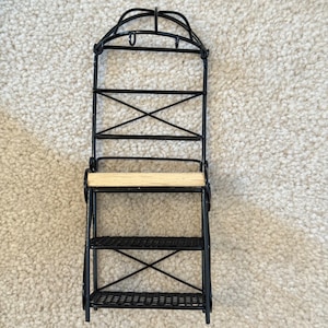 May include: Black metal decorative shelf with three tiers and an arched top. The shelf has a light-colored wooden shelf and is designed for displaying small items. The shelf is suitable for a kitchen or bathroom.