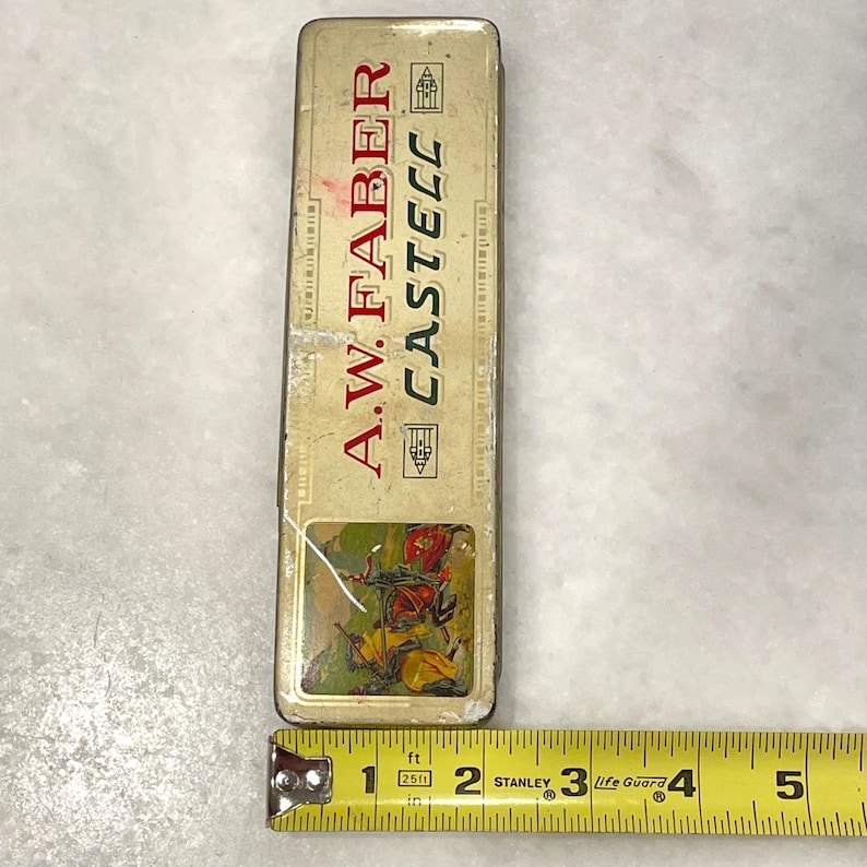 May include: Vintage A.W. Faber Castell pencil case. The rectangular metal case is cream-colored with red lettering and a colorful illustration. The case measures approximately 13 cm long.