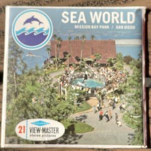 May include: A vintage View-Master reel featuring Sea World in Mission Bay Park, San Diego. The image shows a scenic view of the park with a dolphin logo, a large building, and a crowd of people. The reel is labeled with "21 stereo pictures".