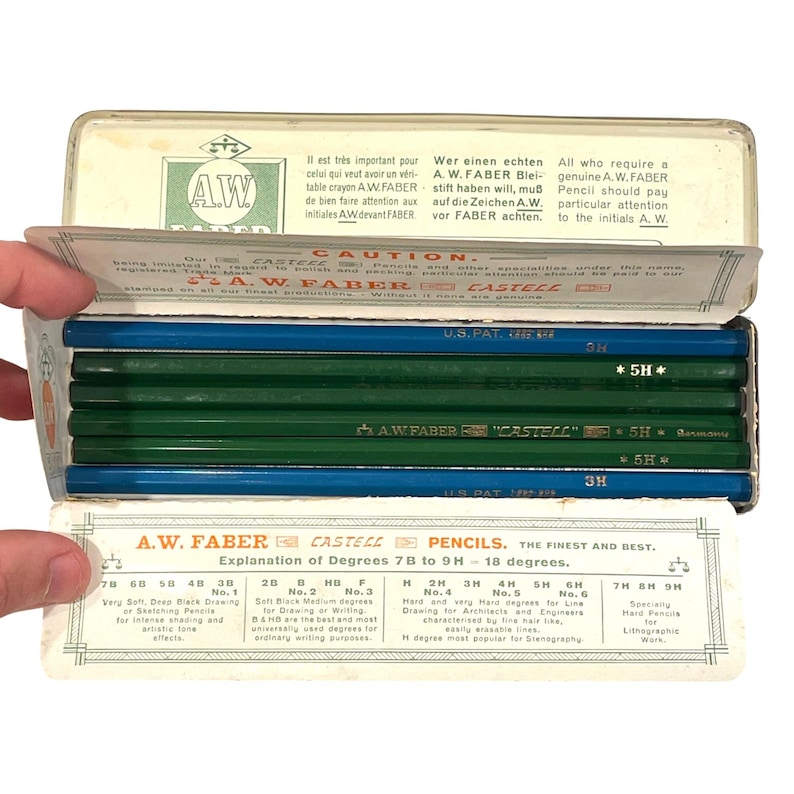 May include: A vintage tin of A.W. Faber Castell pencils. The pencils are green and blue, with degree markings. The tin has text in multiple languages, including "A.W. Faber Castell".