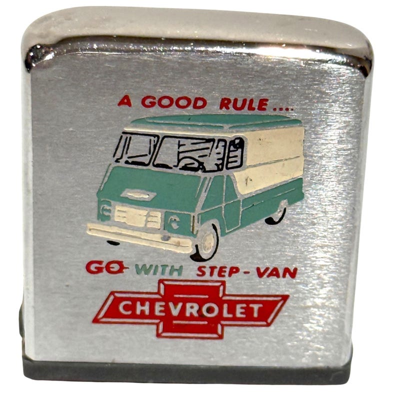 May include: A silver-colored, rectangular lighter with a brushed metal finish. The lighter features a teal and cream-colored step-van graphic with the text "A GOOD RULE... GO WITH STEP-VAN CHEVROLET" in red and black.