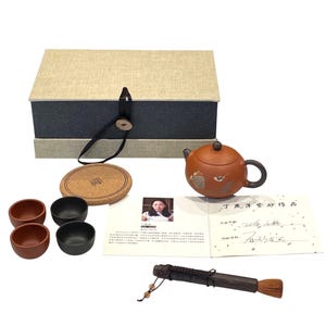 May include: A tea set with a brown teapot, two brown and two black teacups, a cork coaster, and a wooden brush. The set includes a box with a beige lid and a dark blue base. The teapot has a bird design.