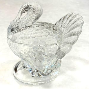 May include: A clear glass turkey-shaped dish with a lid. The dish features detailed feather and body patterns, with a curved neck and head. The base is a round pedestal, and the dish is designed to hold food or decorative items.