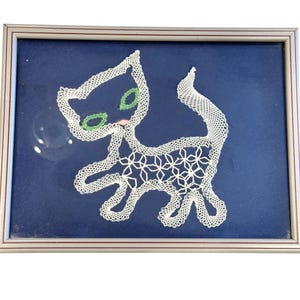 May include: A framed, white lace cat design on a dark blue background. The cat has green eyes and a pink tongue. The frame is silver. The lace cat is a handmade craft.