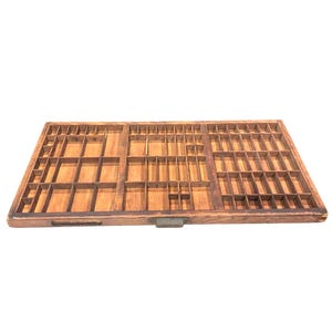 May include: A rectangular wooden printer's drawer featuring a grid of numerous small, open compartments of varying sizes. The tray is constructed from light brown wood and includes a small, centered metal handle on the front edge for easy access.