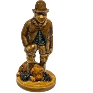 May include: A ceramic figurine of a man in a brown coat and hat, standing on a base with a dog. The figurine has a glossy finish and detailed features. The dog is lying down at the man's feet. The base is round and brown.