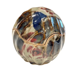 May include: A ceramic lidded jar with a rounded shape and a textured surface. The jar features a mix of colors, including blue, red, and brown, with a glossy finish. The lid fits snugly, creating a decorative storage container.