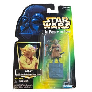 May include: A Star Wars "The Power of the Force" action figure featuring Yoda. The figure is in a clear plastic package with a cardboard backing that includes an image of Darth Vader. The package includes the text "Yoda" and "Jedi Trainer Backpack and Gimer Stick!"