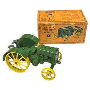 May include: A green and yellow John Deere Model "D" tractor toy with its original box. The tractor has yellow wheels and a green body. The box is orange with text that reads "John Deere Model 'D' Tractor".