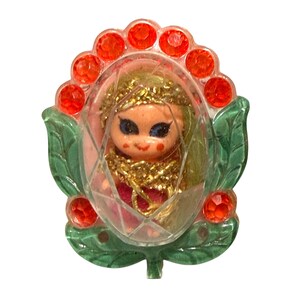 May include: A decorative brooch featuring a small doll with blonde hair and blue eyes encased in a clear, faceted oval plastic frame. The frame is surrounded by green leaf-shaped accents and a series of bright orange circular rhinestones.