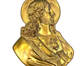 May include: A golden-colored religious bust depicting a man with long hair and a beard, wearing a draped garment. The bust features a halo and a heart emblem. The artwork is likely a religious icon or decorative piece.