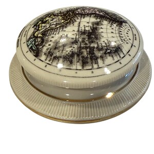 May include: A cream-colored ceramic trinket box with a lid featuring a detailed map of the world. The box has a round base with a ribbed design and a gold trim. The lid is also round and fits snugly on the base.