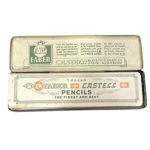 May include: Two vintage pencil boxes, one on top of the other. The top box is white with green text and the words "AW FABER" and "CASTELL". The bottom box is white with the words "AW FABER CASTELL PENCILS THE FINEST AND BEST".