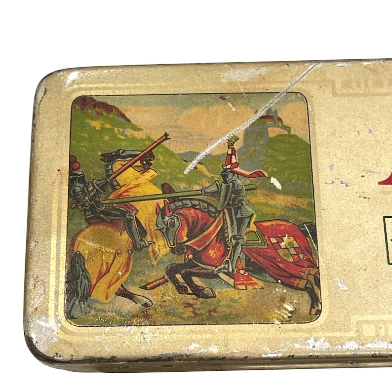May include: A vintage metal box with a gold-coloured exterior. The lid features a colourful illustration of two knights jousting on horseback, set against a backdrop of a landscape with a castle.