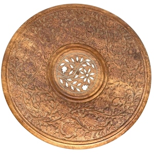 May include: A round, wooden table top with intricate floral carvings. The center features a circular design with white inlay. The detailed carvings cover the entire surface, creating a decorative and ornate piece. The wood has a warm, natural tone.