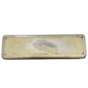 May include: A rectangular metal pencil case with a worn, off-white finish. The case shows signs of wear, with areas of discoloration and a partially peeled sticker. The edges are dark, and the overall look suggests a vintage or antique item.