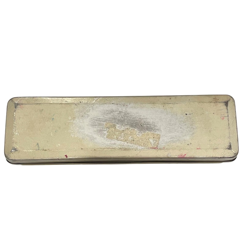 May include: A rectangular metal pencil case with a worn, off-white finish. The case shows signs of wear, with areas of discoloration and a partially peeled sticker. The edges are dark, and the overall look suggests a vintage or antique item.