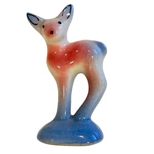May include: A ceramic deer figurine with a glossy finish. The deer has a white head with black accents on the ears and eyes, a red and white body, and blue legs and base. The figurine is a decorative item.