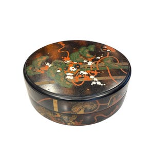 May include: A round, black lacquered box with a lid. The lid features a detailed design with green, orange, and white floral and vine motifs against a speckled background. The box has a layered construction, showcasing traditional Japanese craftsmanship.