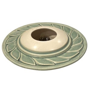 May include: A ceramic flower frog with a light green outer ring and a cream-colored center. The outer ring features a leaf design. The center has a circular opening with metal pins for arranging flowers.
