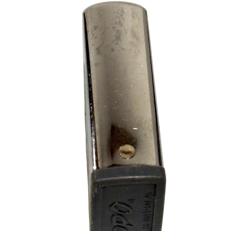 May include: A silver-toned metal lighter with a black base. The lighter has a rectangular shape with rounded edges and a small, circular rivet. The base features the brand name and other text.