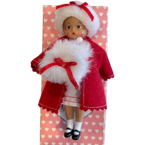 May include: A miniature doll dressed in a red coat, white dress, and a white hat with a red bow. The doll is holding a white muff with a red bow. The doll is standing on a pink box with white hearts.