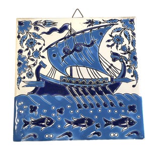 May include: Square ceramic tile with a hand-painted design of a blue ship sailing on blue waves. The tile features floral accents and fish. A metal wire is attached for hanging.
