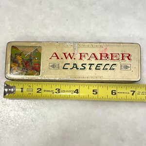 May include: A vintage A.W. Faber-Castell pencil case with a faded gold finish. The case features the brand name in red and green lettering. A colorful illustration of knights on horseback is on the left side. A yellow measuring tape is next to the case.