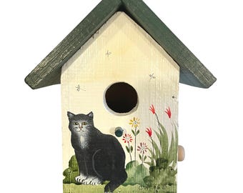 May include: A wooden birdhouse with a cream-colored body and a green roof. The birdhouse features a painted black cat, flowers, and greenery. The birdhouse has a round entry hole and is supported by four wooden feet.