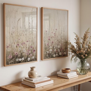 May include: Two framed floral prints with a muted color palette of beige, white, and pink. The prints are hung on a white wall above a wooden table with a vase of dried flowers and books.