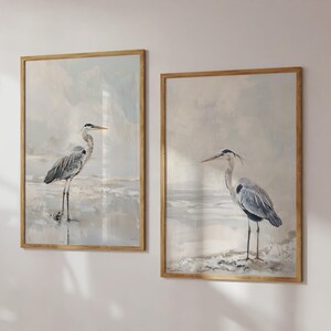May include: Two framed wall art prints featuring grey herons standing in shallow water. Each rectangular print is encased in a light wood frame, showcasing a soft, muted colour palette of grey, white, and beige tones in the background.