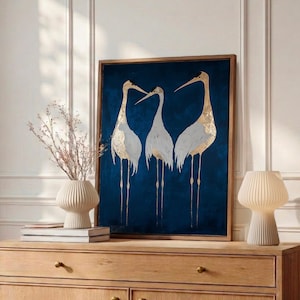 May include: A rectangular framed wall art piece featuring three white and gold cranes standing against a deep navy blue background. The artwork is displayed in a wooden frame and depicts the birds with long legs and pointed beaks.