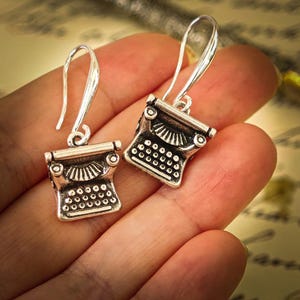 May include: Silver-toned typewriter earrings. Each earring features a miniature typewriter design with detailed keys and a curved hook for pierced ears. The earrings are shown against a soft, blurred background.