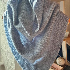 Pode incluir: Um xale triangular feito de fio azul claro e cinza, apresentando um padrão de tricô texturizado em todo o corpo principal e uma borda decorativa recortada ao longo do perímetro. O acessório está drapeado para mostrar sua forma pontiaguda.