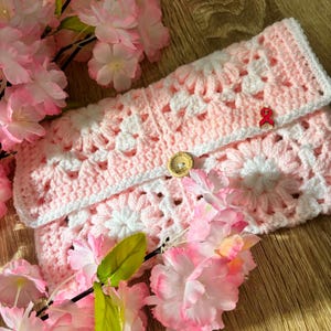 Peut inclure: Une pochette rectangulaire en crochet avec un motif floral de carrés granny roses et blancs. Le rabat est fixé par un bouton en bois rond et comporte une petite épingle à ruban rouge. Les bords sont finis avec une bordure en crochet blanc.