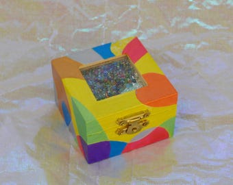 May include: A small, square wooden box with a colorful, abstract design. The box is painted with a mix of bright colors, including yellow, orange, blue, and purple. The lid has a clear window revealing glitter inside. A gold-colored clasp secures the box.