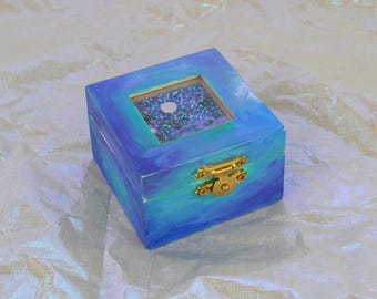 Può includere: Una piccola scatola quadrata di legno dipinta in tonalità di blu e verde acqua con una chiusura color oro. La scatola ha una finestra quadrata trasparente sulla parte superiore, che rivela un interno glitterato. La scatola è su una superficie iridescente strutturata.